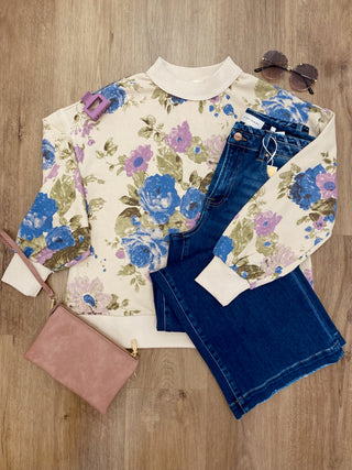 A styled flat-lay outfit on a light wood floor featuring a cream sweatshirt with a blue, purple, and green floral print, folded blue jeans, a dusty pink clutch bag, and sunglasses.