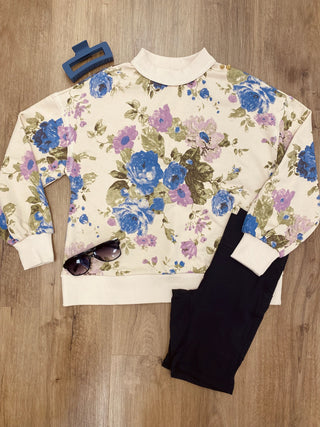 A flat-lay of a cream-colored sweatshirt with a blue and purple floral print, styled with black pants, sunglasses, a blue hair clip, and a wooden floor background.