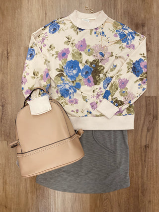 A styled flat-lay outfit featuring a cream sweatshirt with a blue and purple floral pattern, a heather gray skirt, and a beige faux-leather backpack, arranged on a brown wood floor.