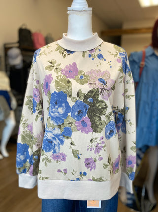 A cream-colored ribbed crewneck sweatshirt with a blue, purple, and green floral print, displayed on a mannequin torso in a clothing store.