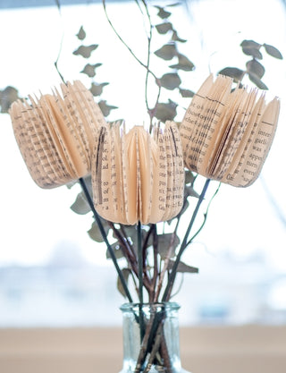Three decorative paper flower bouquets made from folded book pages, arranged in a clear glass vase with dried branches and leaves against a bright, softly blurred background.