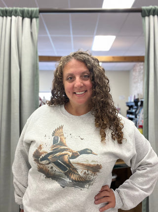 A smiling woman with curly brown hair wearing a light gray sweatshirt with a large graphic of a flying mallard duck on the front, standing in front of a mirror in a retail store.
