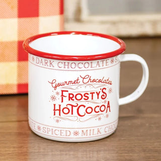 A white enamel-style mug with a red rim and red printed text reading 'Gourmet Chocolates Frosty's Hot Cocoa,' shown on a wooden surface with a blurred background.