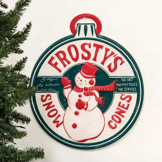 A vintage-style round metal sign for Frosty's Snow Cones, featuring a red and white snowman illustration on a white background with dark green border details. The sign is shown hanging against a white wall with Christmas tree branches visible on the left side.