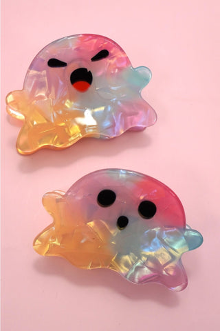 Two translucent ghost-shaped hair clips with an iridescent pastel finish, displayed on a solid pink background. One clip features an angry face, and the other has a surprised face.