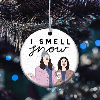 A round white ceramic Christmas ornament hanging from a dark pine tree branch. The ornament features the text 'I SMELL snow' in black lettering above an illustration of two women in winter clothing, with a festive blue bokeh background.