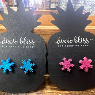 Two black pineapple-shaped display cards on a wooden surface, each holding a pair of glittery snowflake stud earrings. The left card holds bright blue snowflakes and reads 'dixie bliss FOR SENSITIVE EARS'.