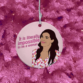 A round hanging ornament with a pink background and printed text reading 'It is literally impossible to be a woman' alongside an illustrated woman, shown hanging on a pink Christmas tree.