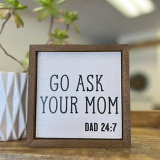A square wooden sign with a white face and black text reading 'GO ASK YOUR MOM DAD 24:7', displayed on a wooden surface with a blurred indoor background and a white planter visible at the left edge.