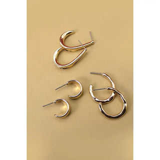A set of four metallic gold earrings arranged on a warm beige background, including two larger abstract hoop-and-arc earrings and two smaller simple hoop earrings.