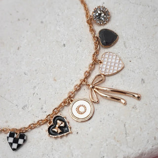 A gold-tone charm bracelet with multiple heart and circular charms, including black enamel hearts, a checkered heart, a bow charm, and a textured heart, displayed on a light stone surface.