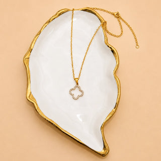 A gold chain necklace with a small flower-shaped pendant set with clear stones, displayed on a white and gold ceramic tray against a warm beige background.