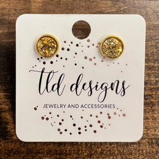 A pair of round stud earrings with gold-toned metal and glittery gold centers, displayed on a white jewelry card that reads 'ltd designs' and 'JEWELRY AND ACCESSORIES,' placed on a wooden surface.