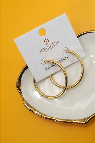 A pair of gold hoop earrings on a white display card labeled JOSSLYN 18K GOLD DIPPED, placed on a white plate with a gold rim against a bright yellow background.