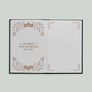 An open notebook or journal with a black cover, showing two white interior pages. The left page has a brown floral border and the text 'A MOMENT I WAS SO PROUD OF YOU.
