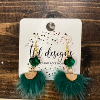 A pair of dangling earrings displayed on a white card against a rustic dark wood background. The earrings have gold-tone hooks, green faceted beads, gold-tone semicircle charms, and teal faux fur tassels.