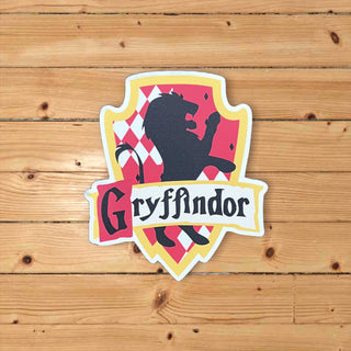 A die-cut sticker of the Gryffindor crest from Harry Potter, featuring a black lion silhouette over a red and white harlequin pattern shield, with a white banner reading 'Gryffindor' below, displayed on a light brown wooden plank background.