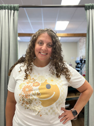 A smiling woman with curly brown hair wearing a white graphic t-shirt with a gold smiley face and floral design, standing in front of a fitting room curtain.