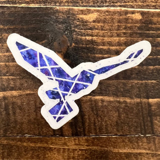 A die-cut sticker of a stylized bird with spread wings, filled with a blue and purple galaxy pattern and outlined in white, shown on a rustic dark brown wooden surface.