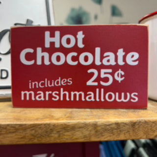 A red rectangular sign with white text reading 'Hot Chocolate includes 25¢ marshmallows' sitting on a wooden surface.