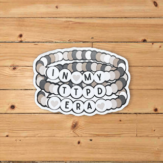 A die-cut sticker featuring a stacked coiled design with the text 'IN MY TTPD ERA' in black letters on white circles, separated by gray hearts. The sticker is placed on a light brown wooden plank background.