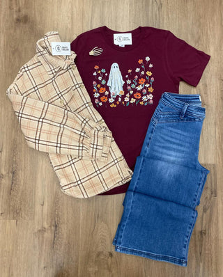 A flat-lay of a three-piece children's outfit on a light wood floor, featuring a maroon t-shirt with a ghost and floral graphic, a beige plaid button-up shirt, and blue jeans.