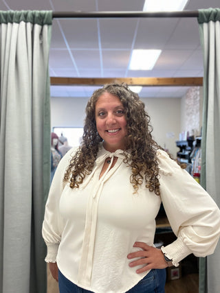 A woman with curly brown hair wearing a cream-colored long-sleeve blouse with a tie-neck detail, standing in front of a mirror in a fitting room with gray curtains.