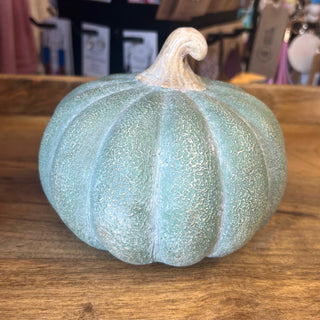 A decorative faux pumpkin in a distressed light blue-green color with a textured, cracked finish and a natural beige stem, sitting on a wooden tabletop.