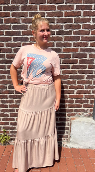 Woman wearing a pink t-shirt with a graphic design and a beige skirt standing against a brick wall.
