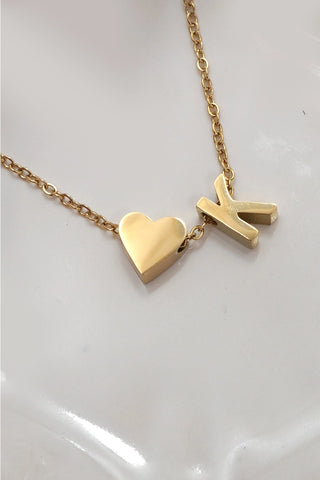 A gold chain necklace featuring a polished gold heart pendant and a matching gold block letter 'K' pendant, displayed on a glossy white surface.