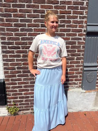 Person wearing a graphic t-shirt and light blue skirt standing against a brick wall.