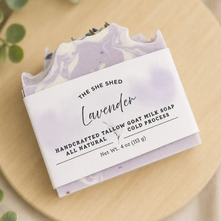 A lavender goat milk soap bar wrapped in a white paper band labeled 'THE SHE SHED Lavender,' photographed on a light wood surface with soft green leaves at the edge.