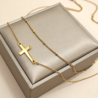 A gold cross pendant necklace draped over a gray satin jewelry box on a light beige background.