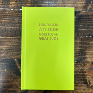A bright lime green hardcover notebook with the text 'LESS FUCKIN ATTITUDE MORE FUCKIN GRATITUDE' printed in a muted gold-gray font on the front cover, displayed flat on a dark wood background.