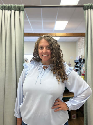 A woman with curly brown hair wearing a light blue long-sleeve blouse with ruffled shoulders and a tie-neck detail, standing in a fitting room with green curtains.