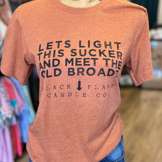 A heathered terracotta orange t-shirt with black graphic text reading 'LETS LIGHT THIS SUCKER AND MEET THE OLD BROADS BLACK FLAME CANDLE CO.' displayed on a mannequin torso in a retail store.