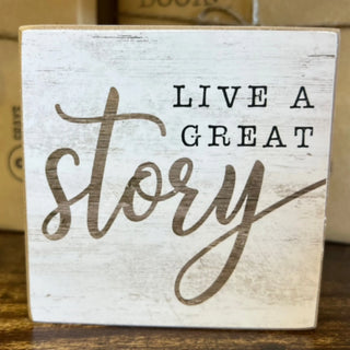 A square wooden sign with a distressed white finish and the phrase 'LIVE A GREAT story' in dark brown lettering, displayed on a wooden surface.