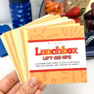 A hand holds a fan of 'Lunchbox Lift-Me-Ups' affirmation cards, with a blue water bottle, strawberries, and grapes blurred in the background.