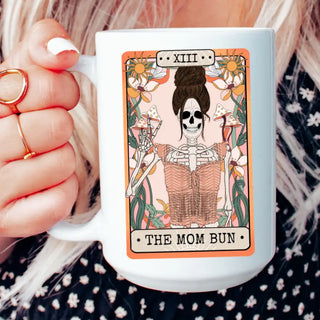 A white ceramic mug featuring a tarot card illustration of 'The Mom Bun' with a skeleton in a bun, held by a person with blonde hair wearing a black and white floral top.