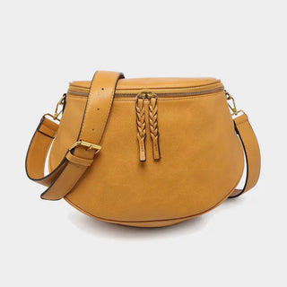 A mustard yellow leather crescent-shaped crossbody bag with a top zipper, braided leather zipper pull, and an adjustable shoulder strap, shown on a light gray background.