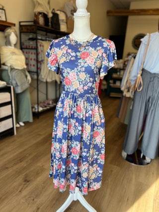 A short-sleeve midi dress with a blue floral print featuring pink, red, and white flowers, displayed on a white mannequin in a clothing boutique.