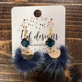 A pair of bohemian-style dangle earrings displayed on a white card. The earrings feature gold hooks, dark teal faceted beads, gold crescent moon charms, and fluffy navy blue faux fur tassels.
