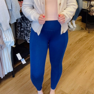 A person standing in a clothing store wearing bright royal blue leggings, a light pink top, and a cream cardigan. The background shows a wooden floor and clothing racks.
