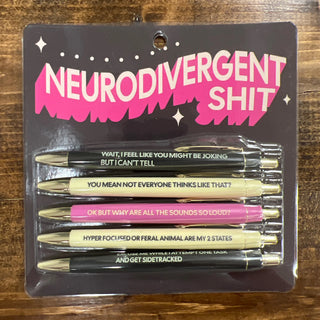A set of five novelty pens in a blister pack on a dark brown wooden surface. The packaging card features the text 'NEURODIVERGENT SHIT' in white and pink.