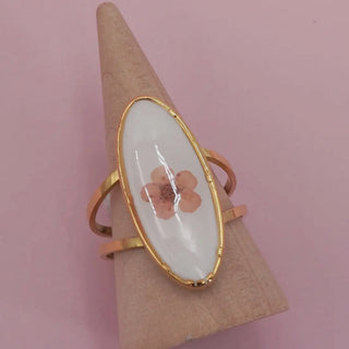 A gold ring with an oval white inlay featuring a pressed pink flower, displayed on a cone stand against a solid pink background.