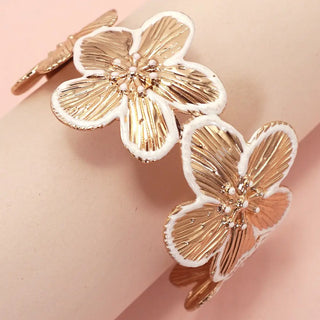 A close-up of a floral cuff bracelet with three large gold-tone flower charms outlined in white, worn on a pale pink wrist against a soft pink background.