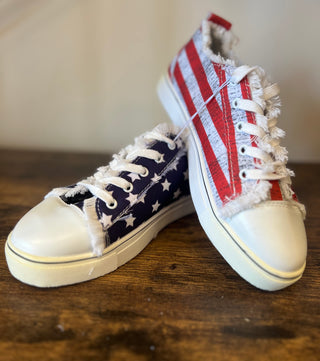 A pair of distressed canvas sneakers with an American flag theme, featuring a navy blue base with white stars on one shoe and red and white stripes on the other, resting on a wooden surface.