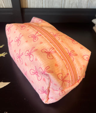 A peach-colored zippered pouch with a pink bow pattern, resting on a dark wooden surface.