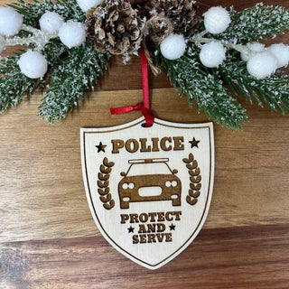 A wooden police badge ornament hanging from a red ribbon, displayed on a wooden surface with festive greenery and white pom-pom berries above it.