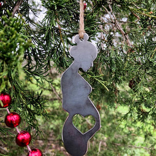 A dark gray metal Christmas ornament shaped like a pregnant woman with a heart-shaped belly cutout, hanging from a tree branch with red ornaments in the background.
